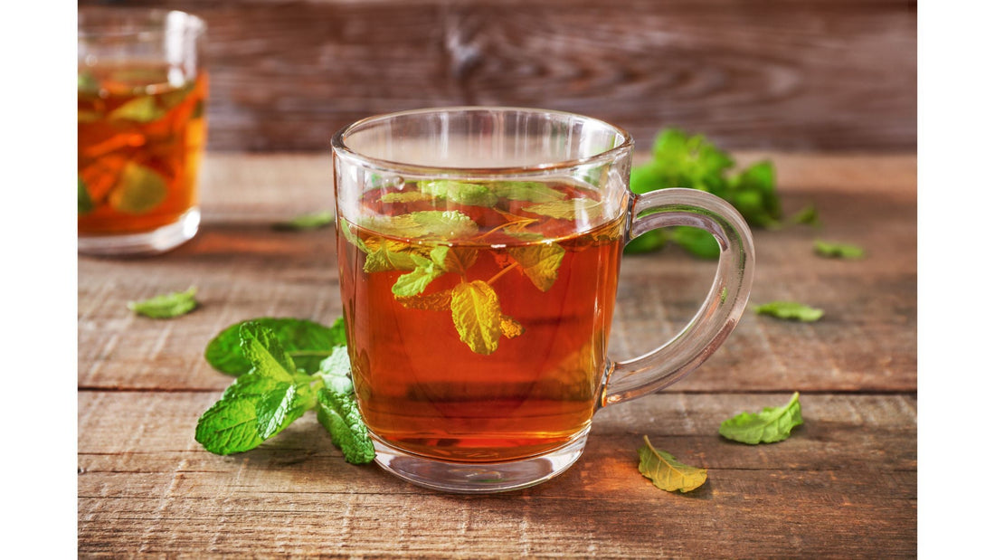A clear glass mug filled with tulsi tea and fresh green tulsi leaves, sitting on a rustic wooden surface.