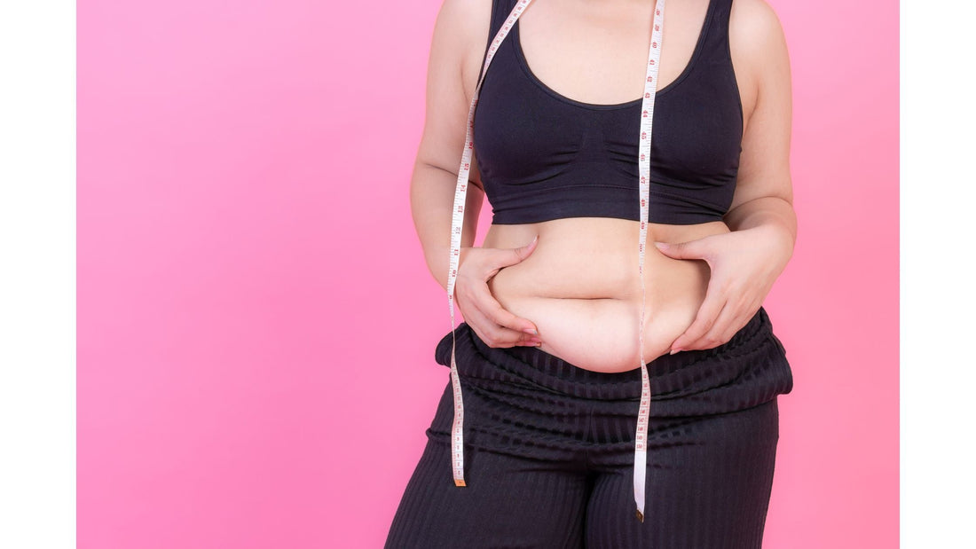 A woman in a black sports bra and pants pinches excess fat around her stomach while a measuring tape hangs around her neck, against a pink background.