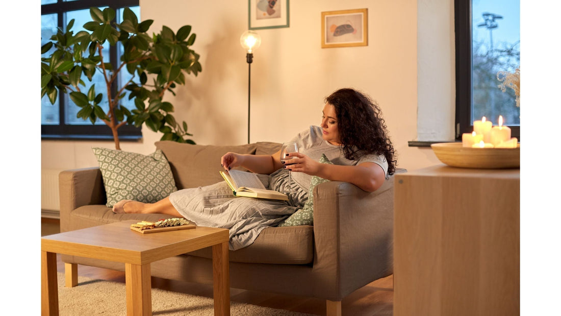 A woman with curly hair sitting on a brown couch, reading a book and holding a glass, in a cozy living room setting with plants and soft lighting.